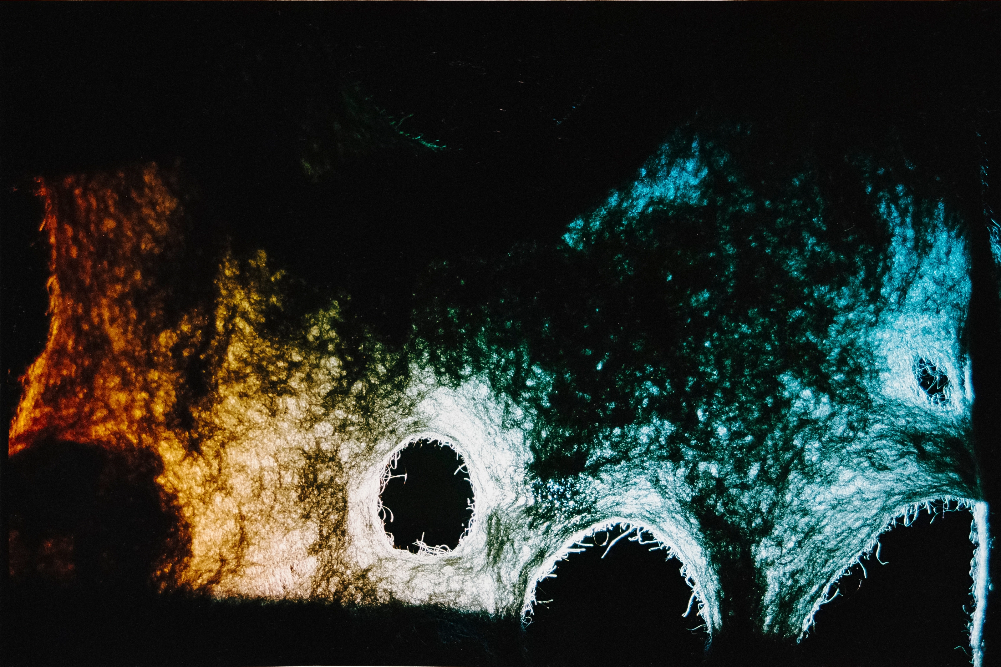 Dramatically lit felted-wool sculpture with holes, in a warm orange-to-teal gradient against a dark background.