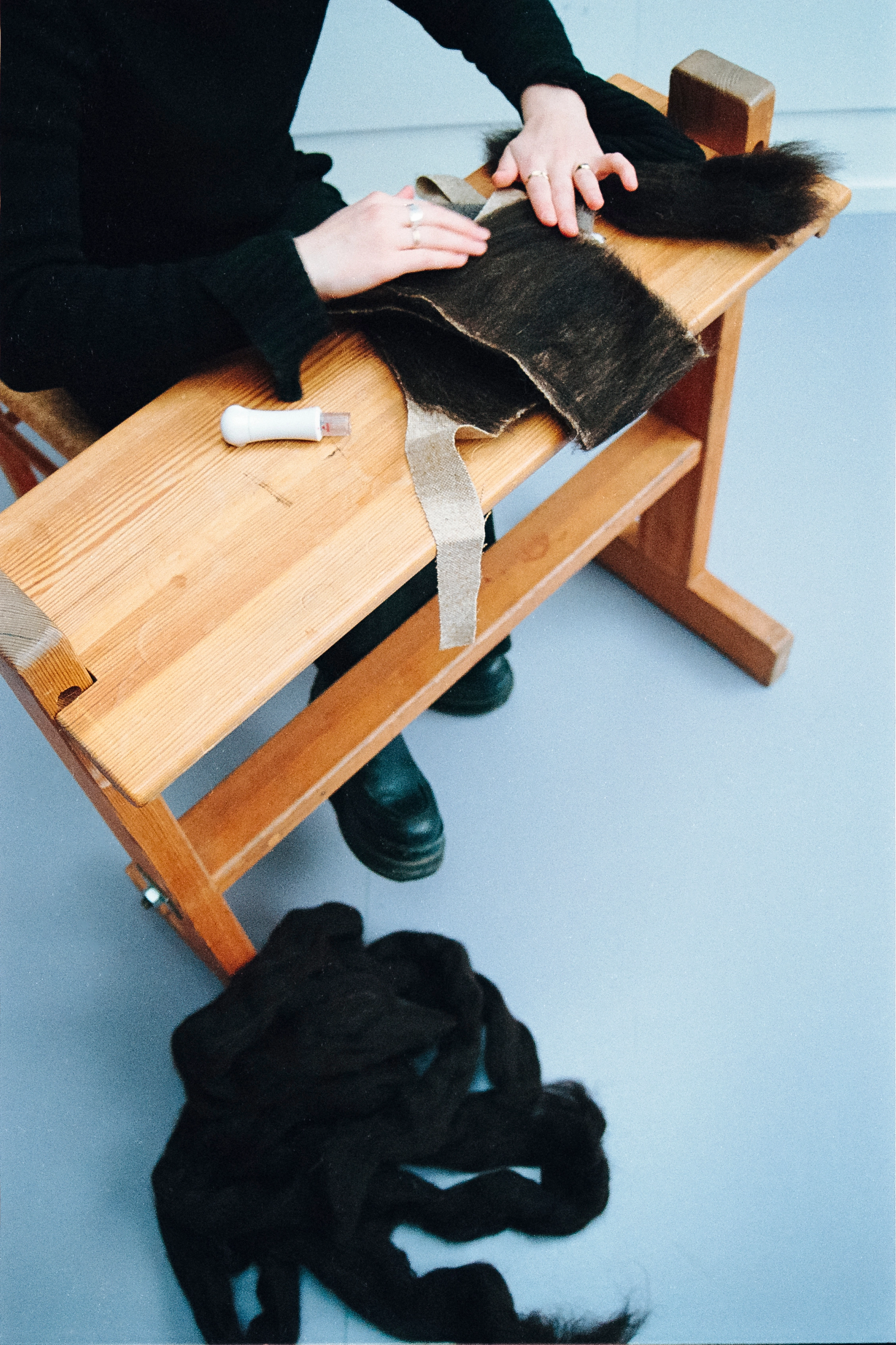 Analog photograph of someone working dark wool fibers on a wooden bench, with carded wool lying on the floor.