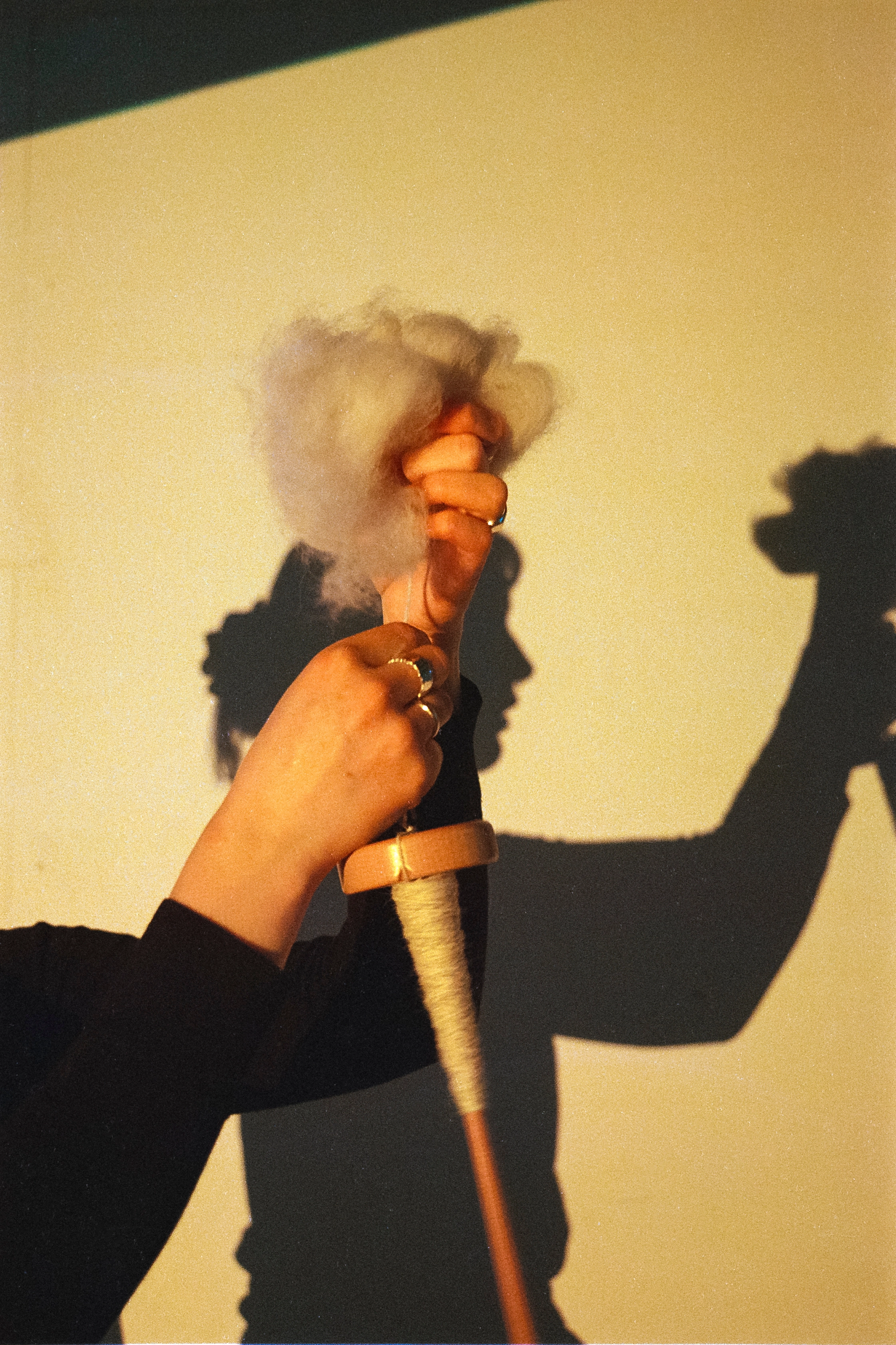 Analog photograph of a hand spinning raw wool onto a spindle, with the photographer's shadow visible on the wall behind.