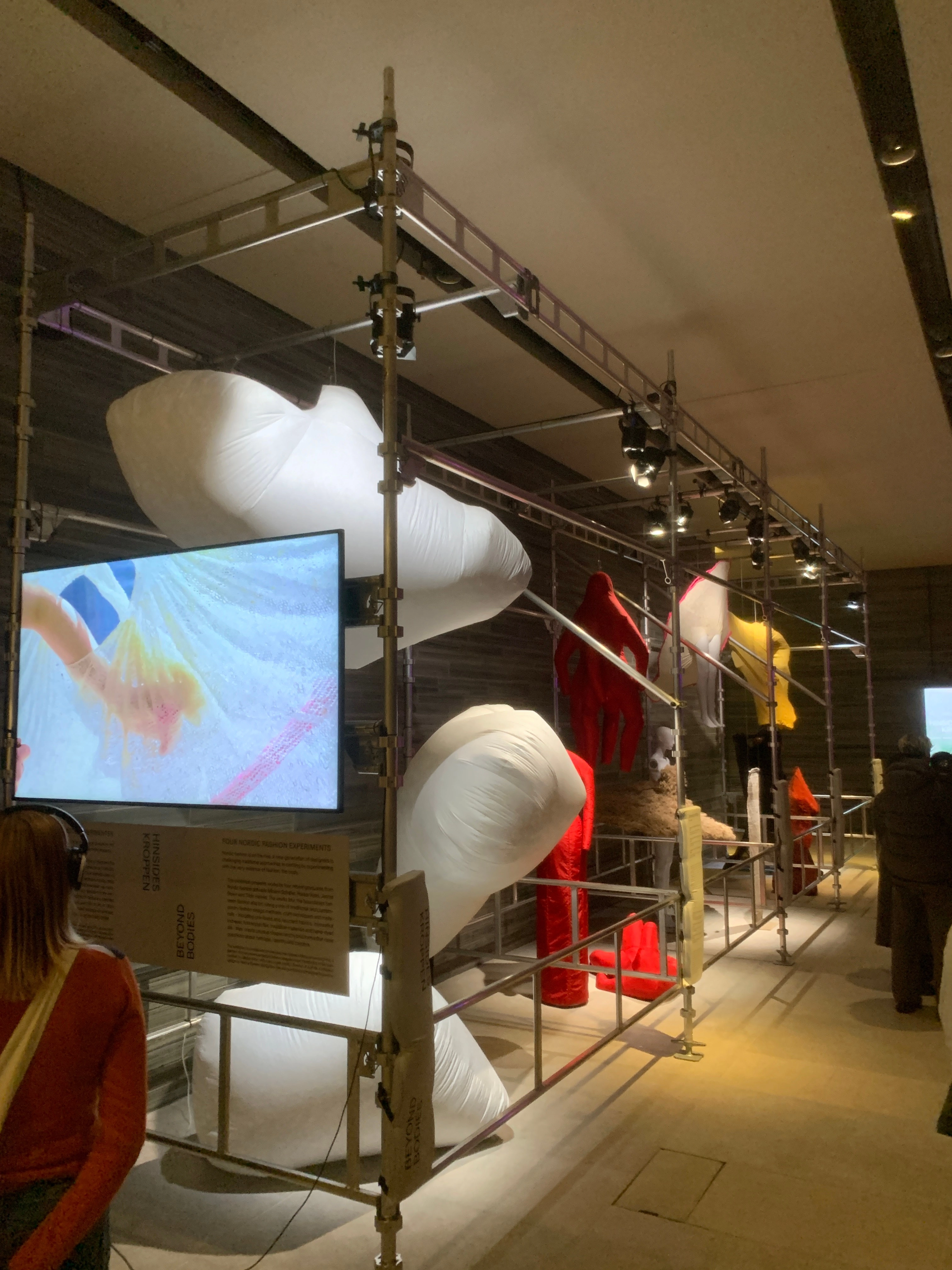 Photograph from the opening night of the finished exhibition — white, red, and yellow textile forms in the scaffolding structure, with the "Hinsides kroppen / Beyond Bodies" panel and a screen visible.